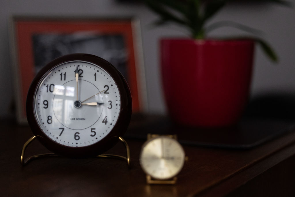 A tabletop with an analog clock in focus and a wristwatch in the foreground, slightly out of focus. A potted plant is to the right