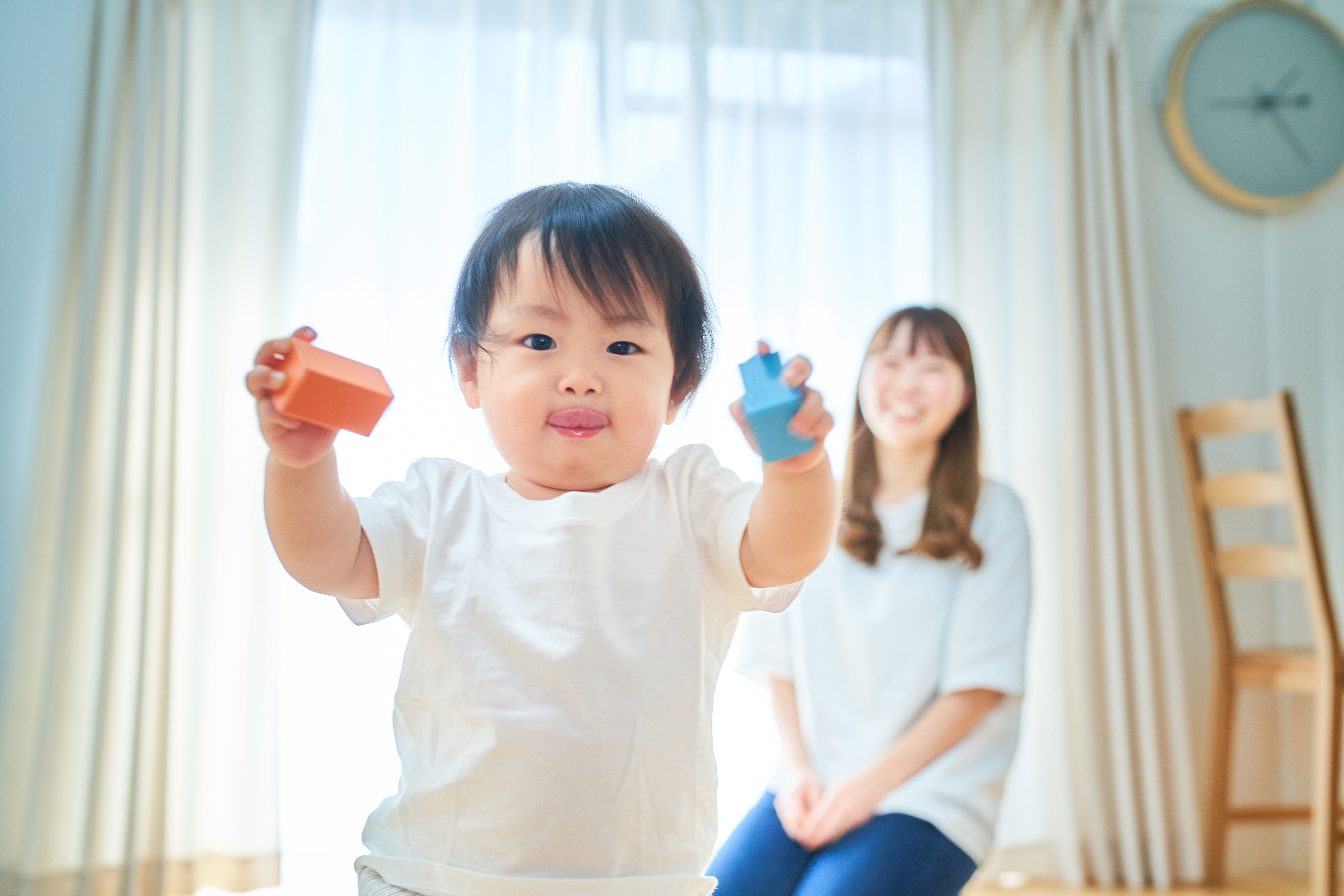 Toddler holding toys with smiling woman seated in the background