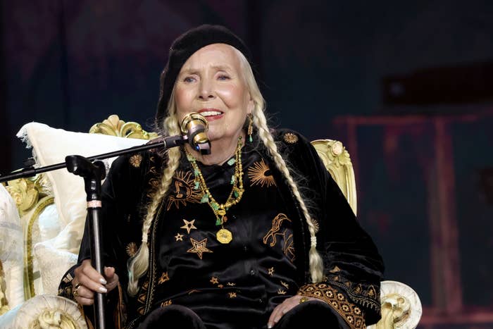 Joni Mitchell seated with microphone, dressed in black with star motifs, at a music event