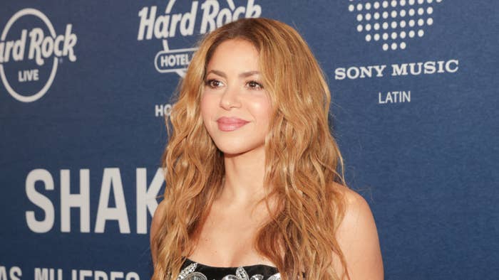 Shakira on a backdrop, wearing a strapless outfit with a detailed pattern, smiling at an event