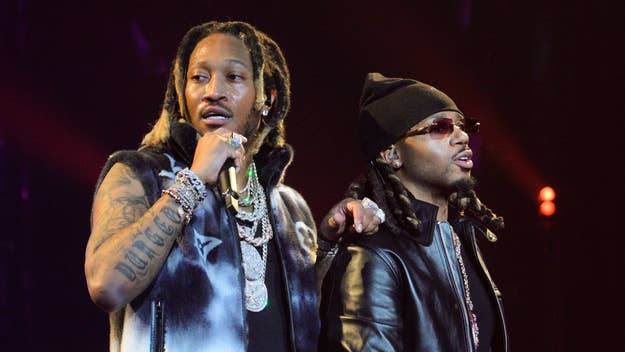 Two hip-hop artists performing on stage, one holding a microphone. They wear stylish jewelry and sunglasses