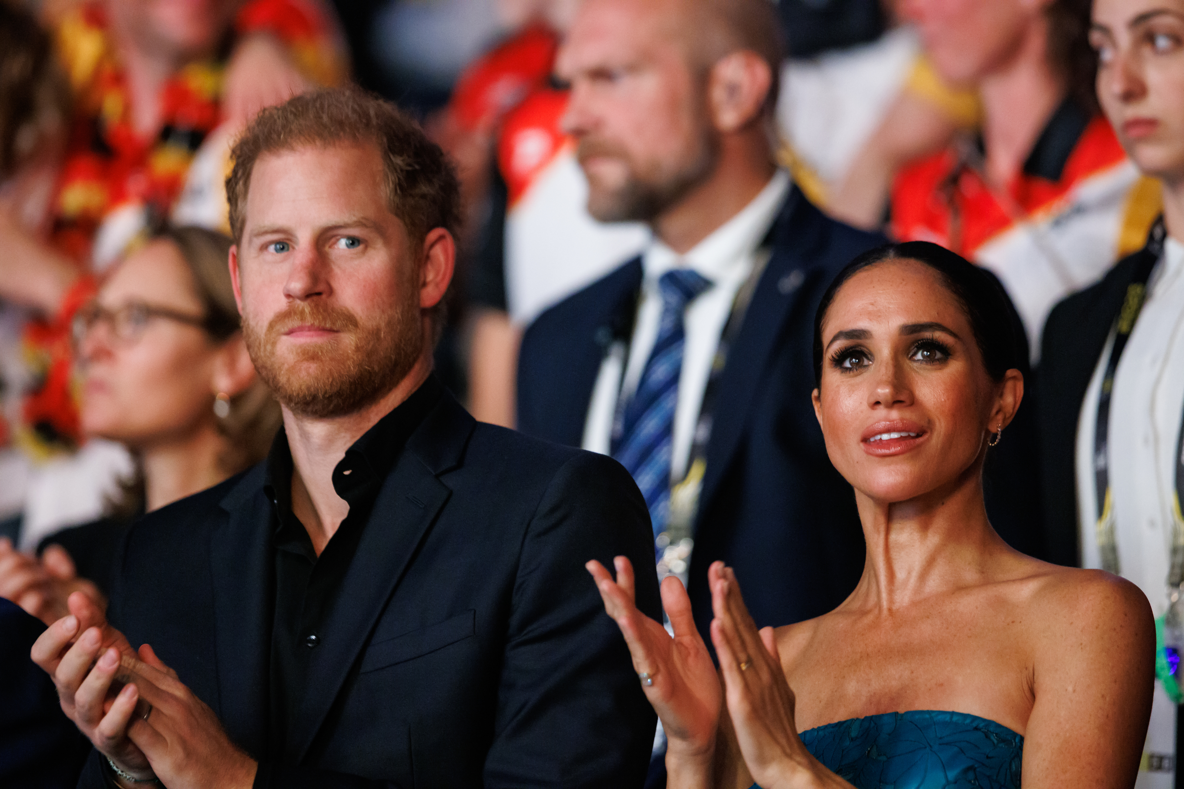 Prince Harry and Meghan Markle seated, clapping, Meghan in a strapless top