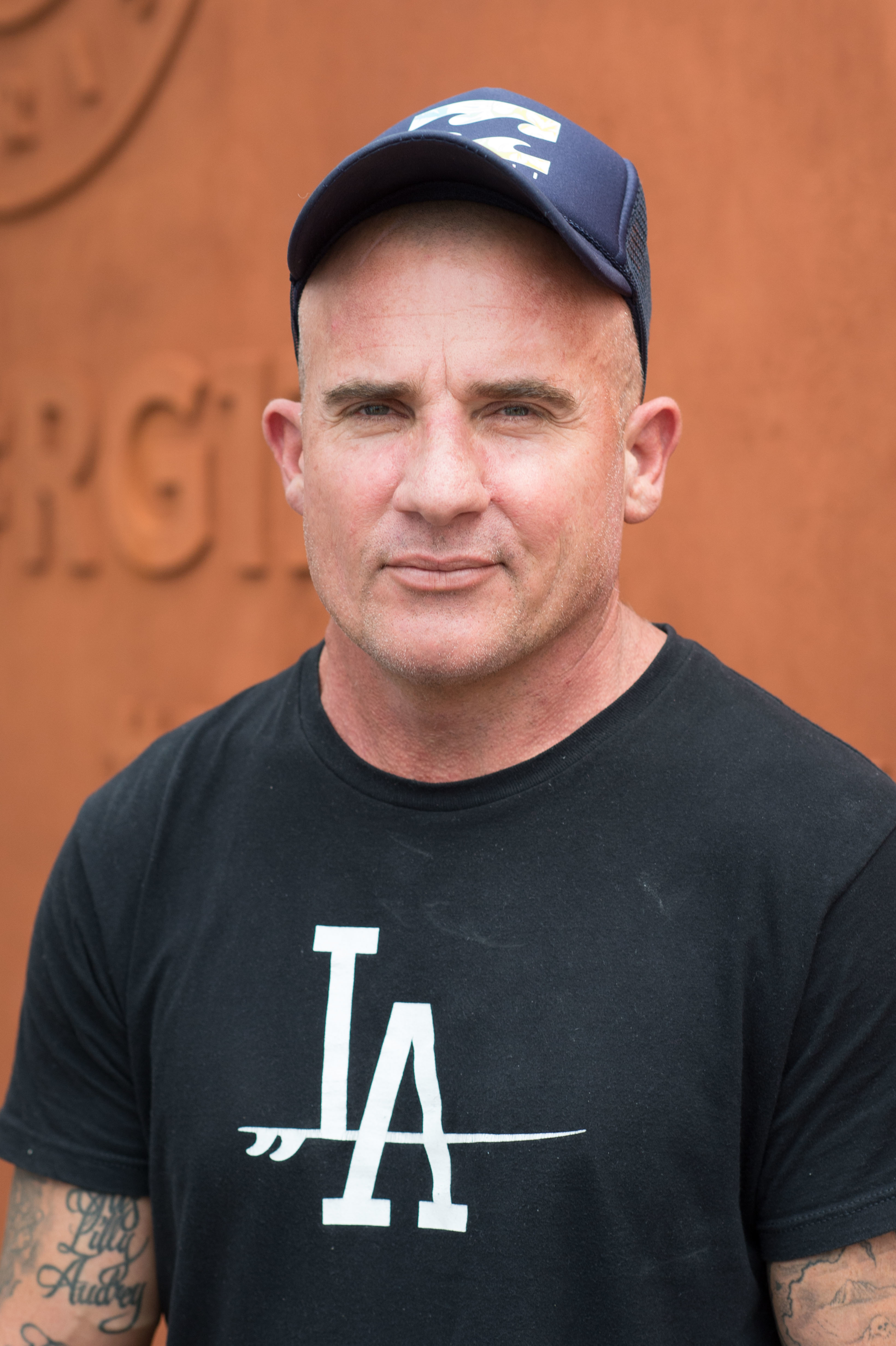 Dominic Purcell in a black t-shirt and cap, standing in front of a textured backdrop