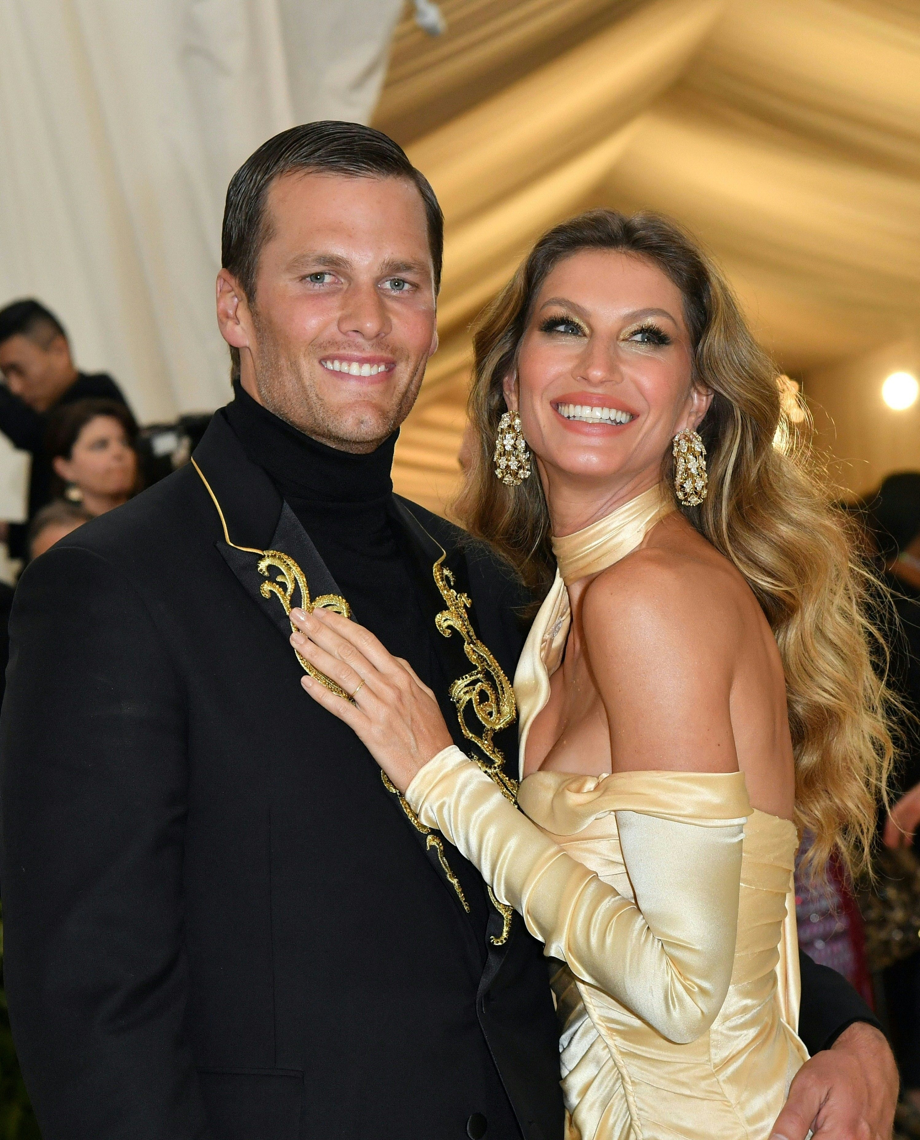 Tom Brady and Gisele Bündchen smiling at an event, Brady in a black outfit with gold detailing, Bündchen in an off-the-shoulder gold gown