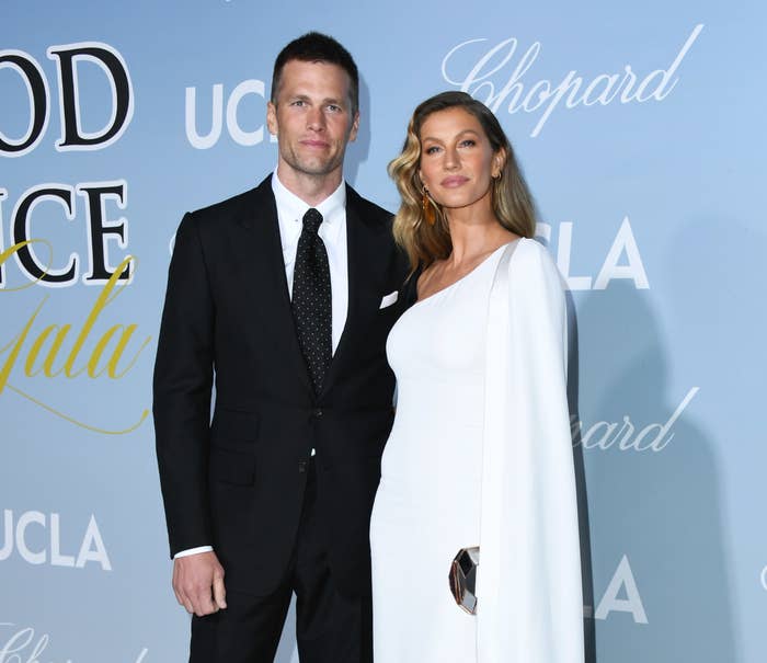 Tom in a black suit and tie and Gisele in a white dress with a draped sleeve at a media event