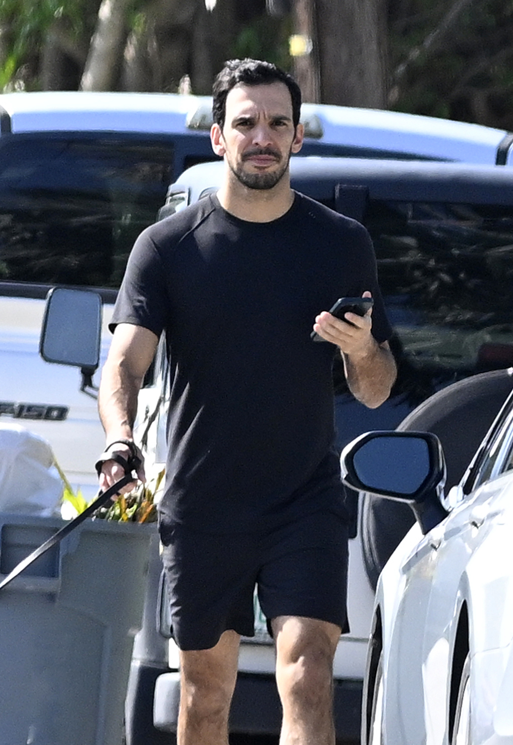 Joaquim in casual athleticwear holding a phone and walking by a car