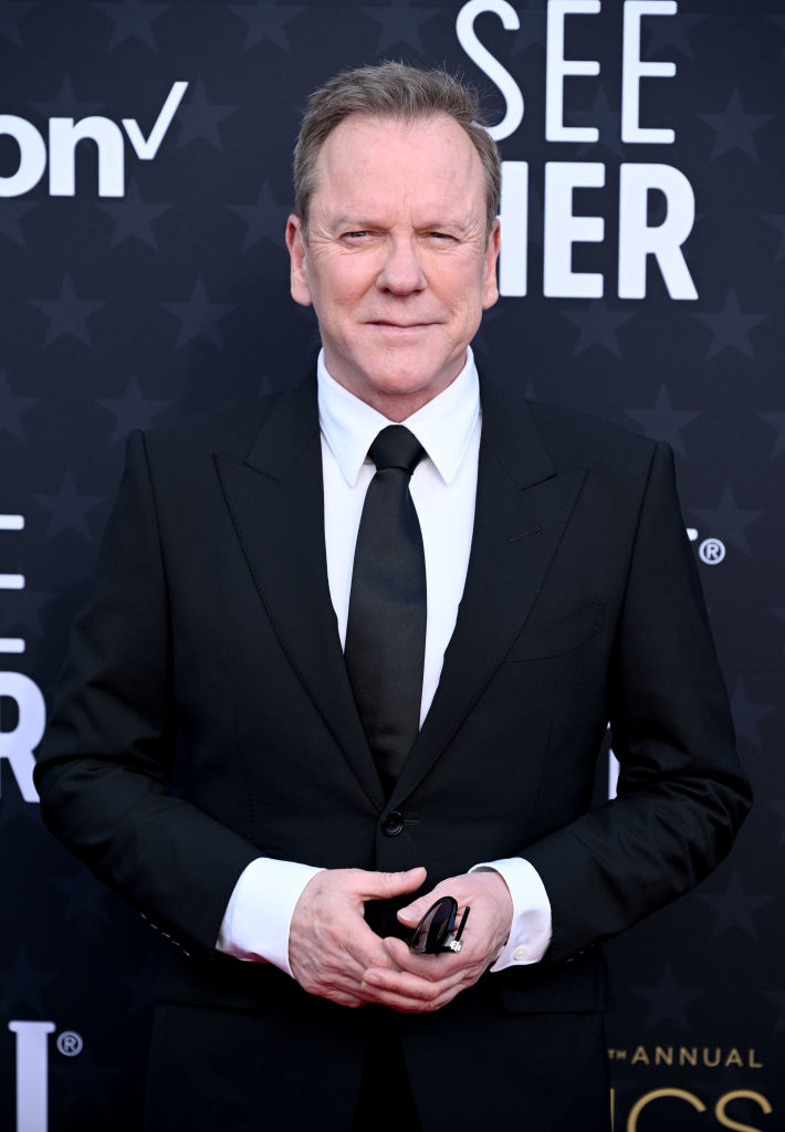 Kiefer in a classic black suit and tie poses on the event red carpet