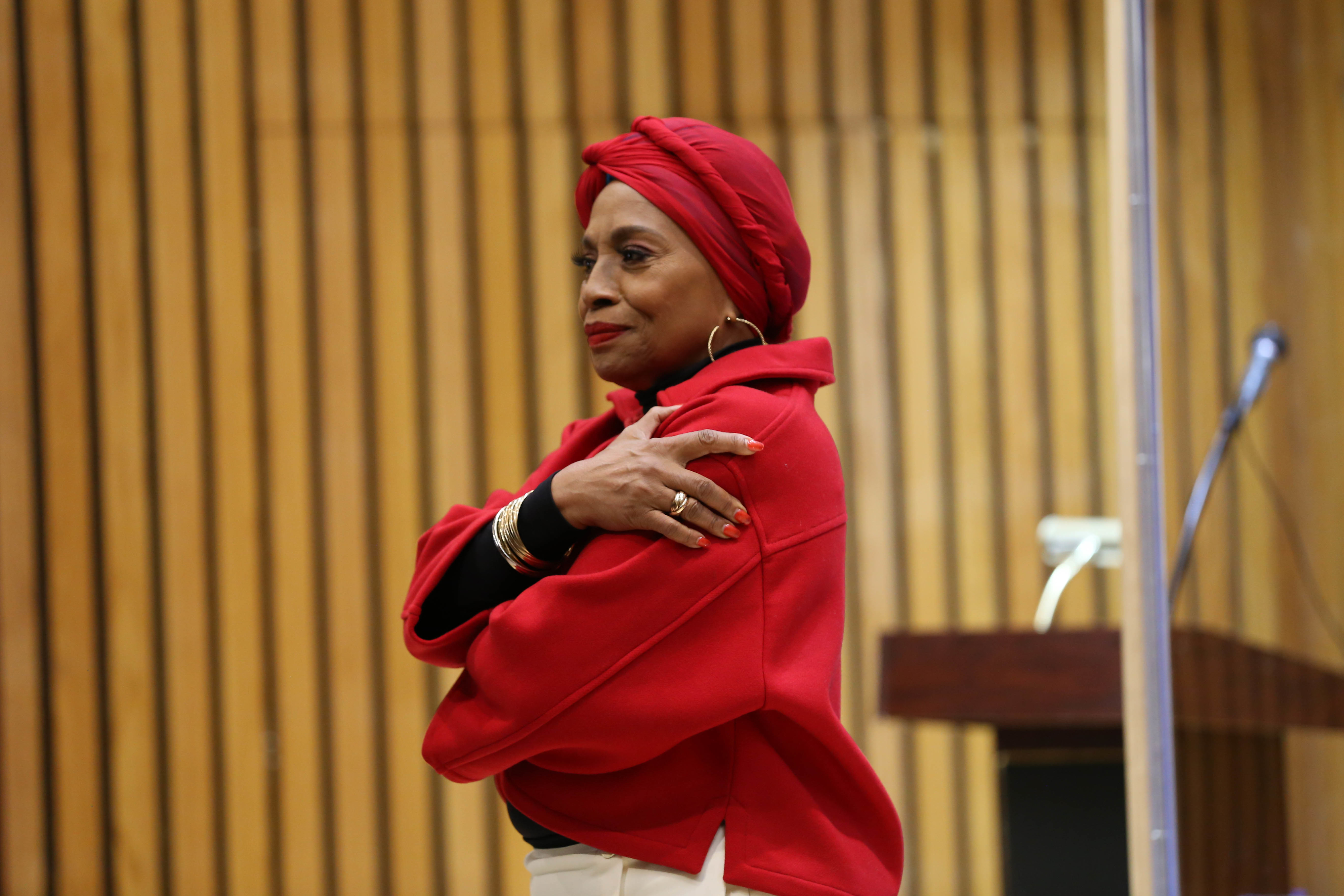 Jenifer in a headwrap and top with arms crossed standing by a podium