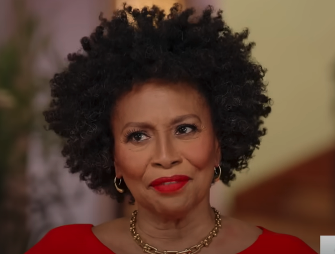 Close-up of Jenifer Lewis wearing a red top and gold necklace, with a warm smile
