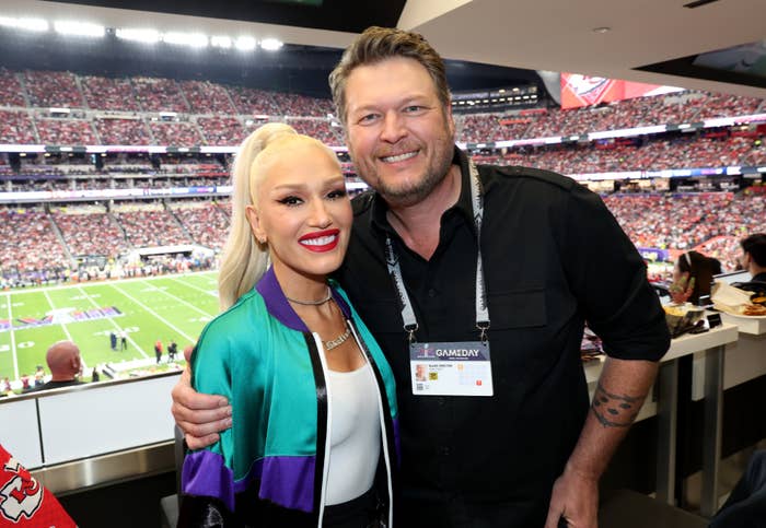 Gwen Stefani in a jacket smiling with Blake Shelton, in a black shirt, at a sports event