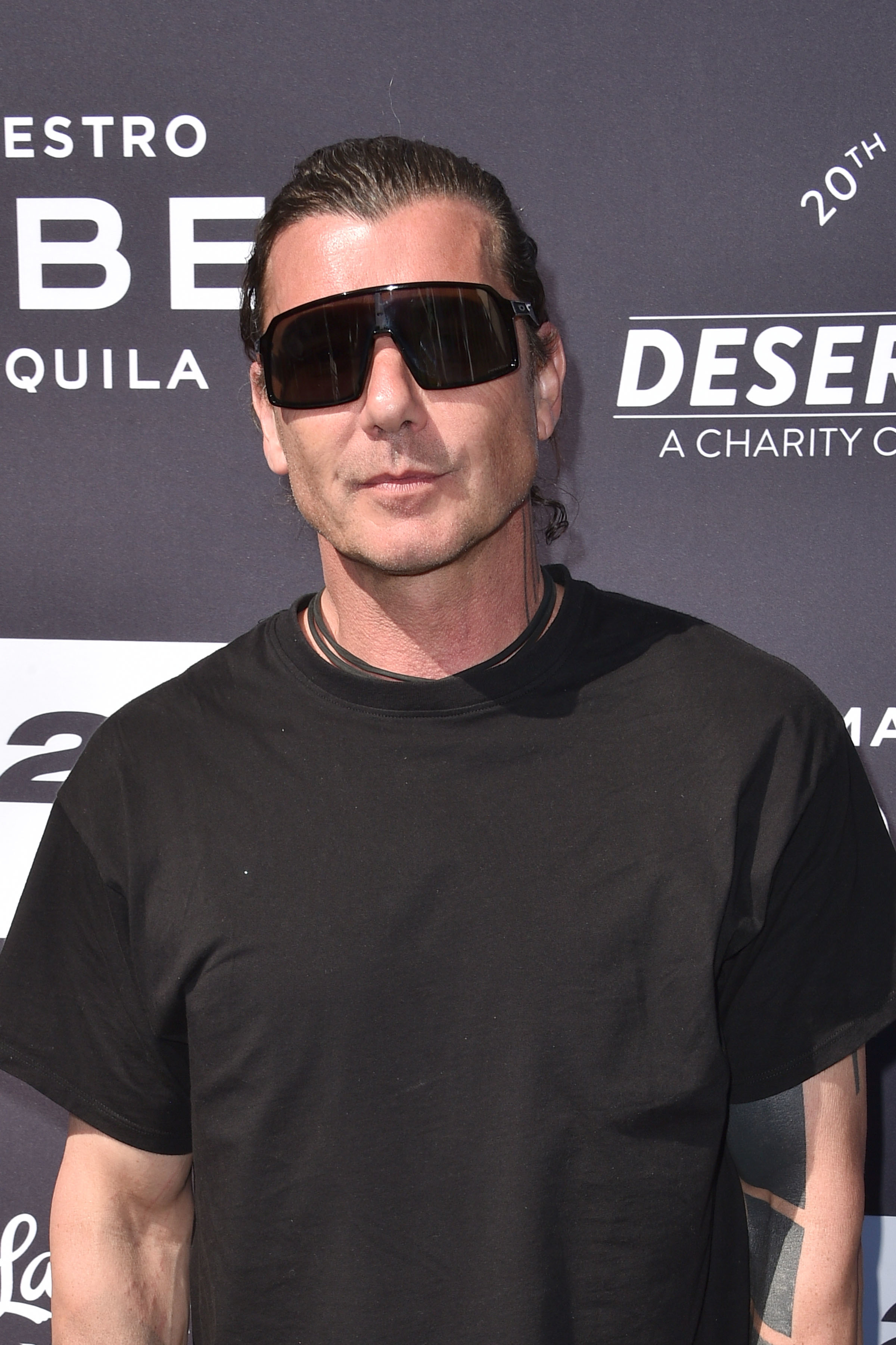Gavin Rossdale stands with a neutral expression, wearing a black T-shirt and dark sunglasses