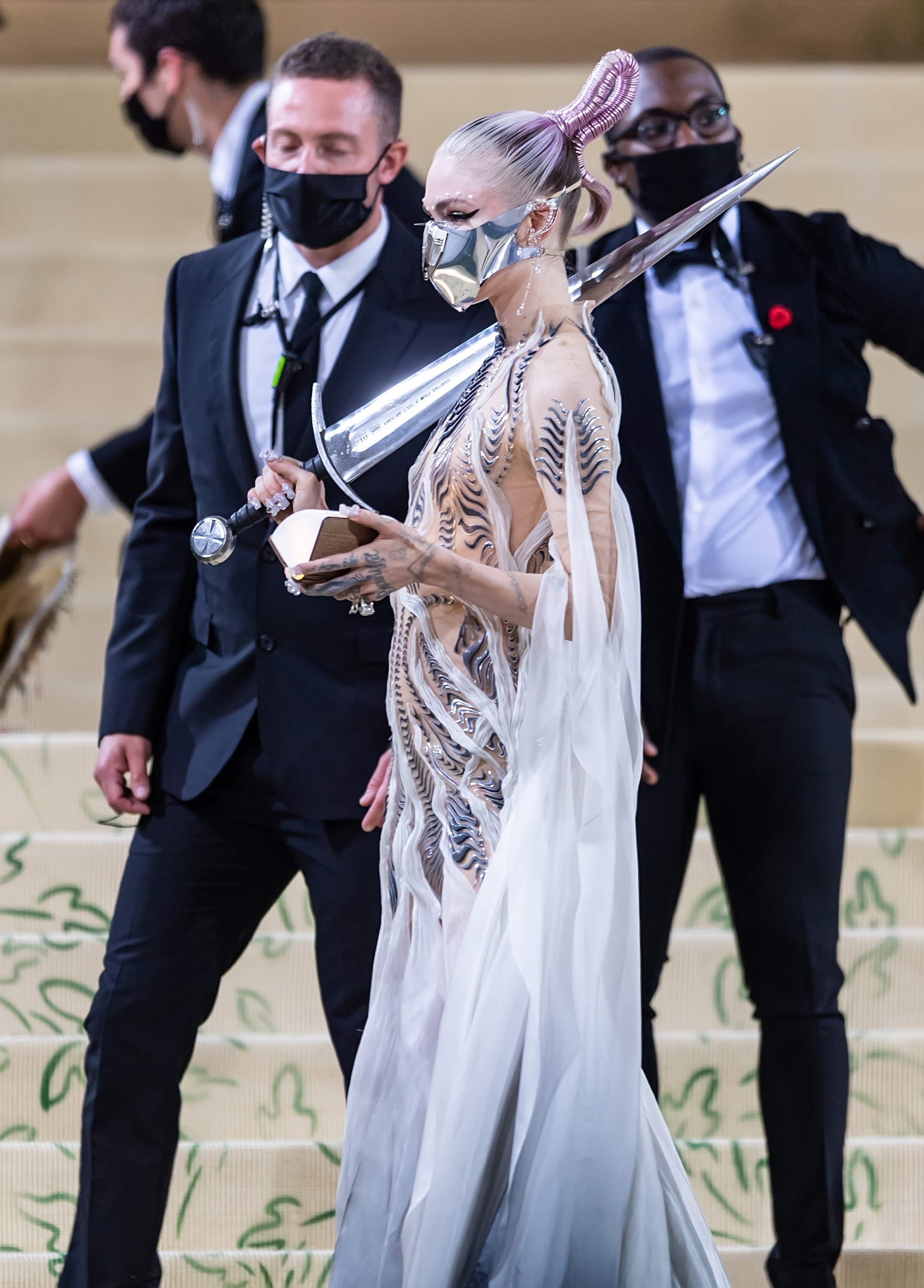 Grimes in a futuristic, sculptural gown with metallic accents, at a formal event, with masked attendees in the background