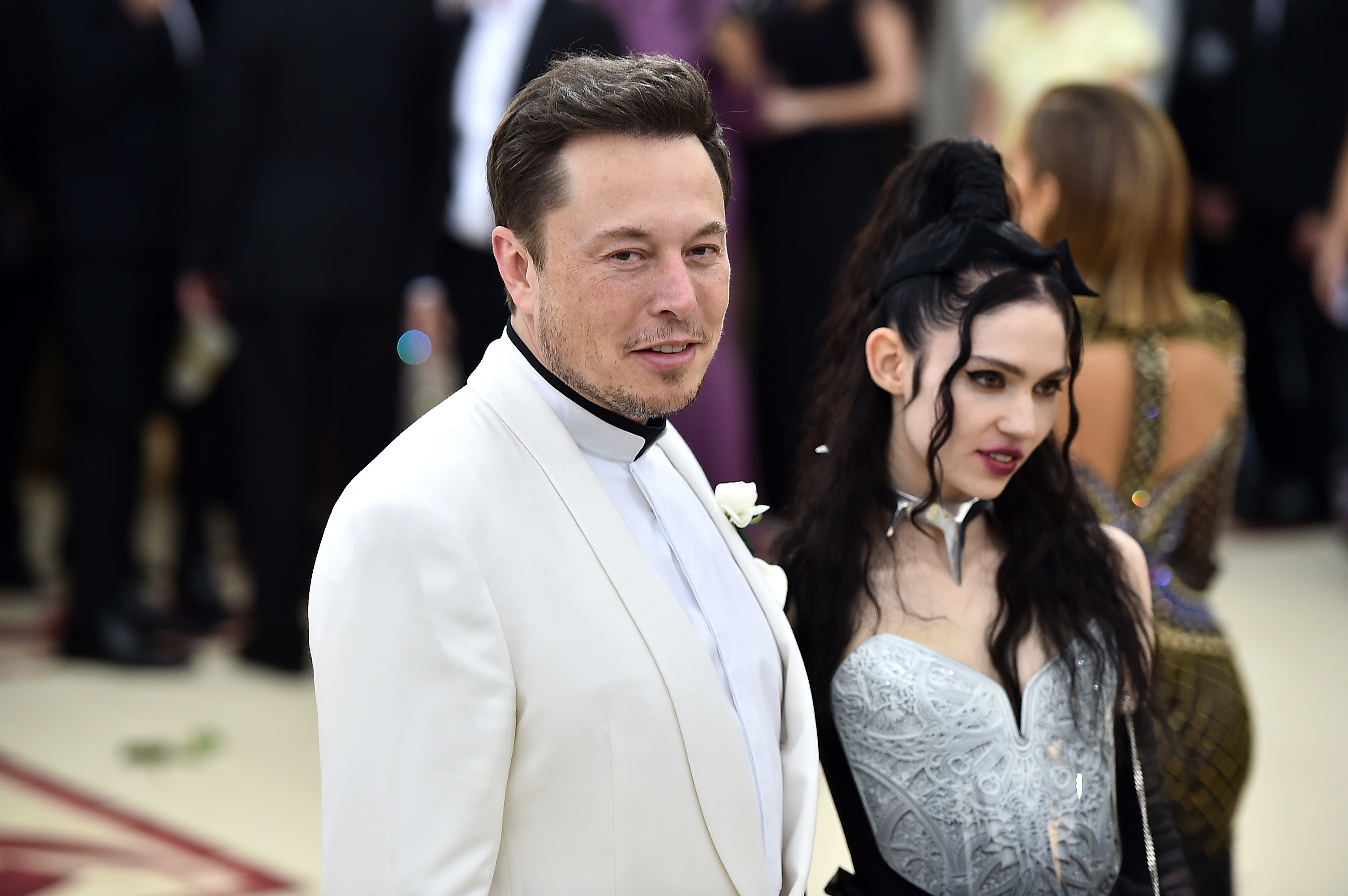 Elon Musk in a tuxedo and a Grimes in a silver outfit with structured shoulders and a choker at an event