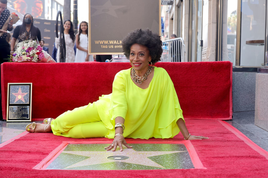 Jenifer lying on her side on the red carpet beside her star plaque on the Hollywood Walk of Fame, wearing a bright outfit and smiling