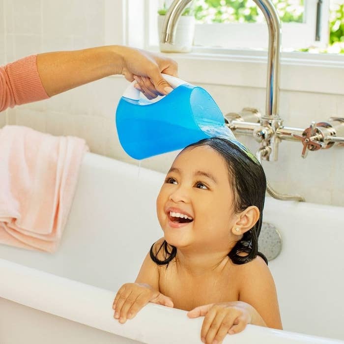 Adult pouring water from blue pitcher over smiling child&#x27;s head in a bathtub, demonstrating hair washing