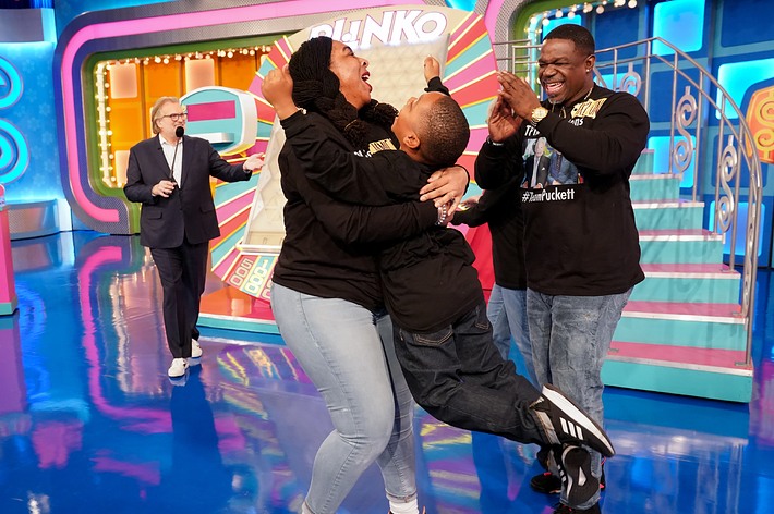 Three people celebrating on a game show stage with a host in the background