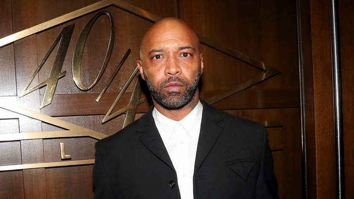 Joe Budden in a black suit with a lapel pin, standing before a wooden backdrop with &#x27;40/40 Club&#x27; etched in it