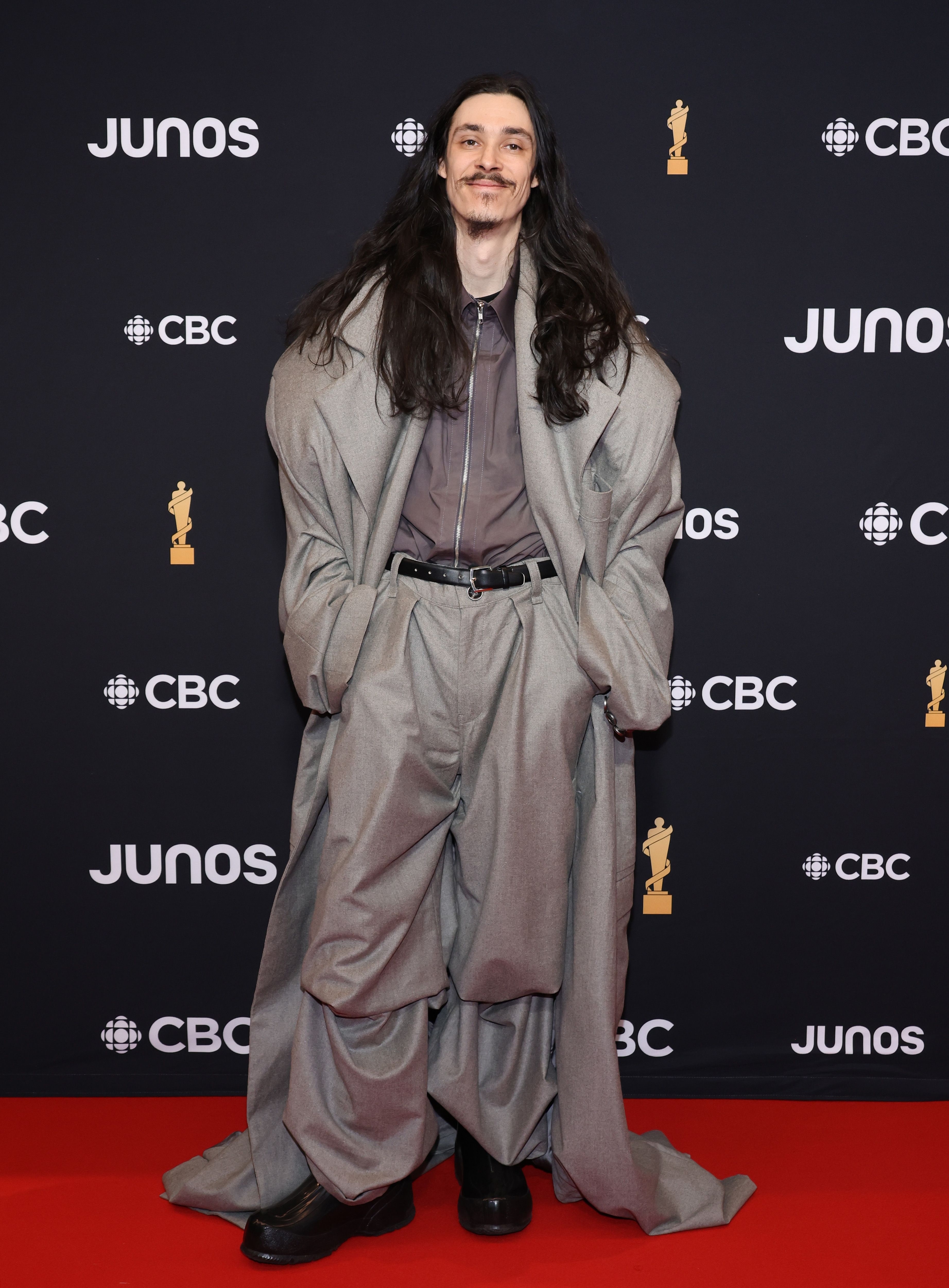 Person in oversized gray suit and long coat poses at event with &#x27;JUNOS CBC&#x27; backdrop