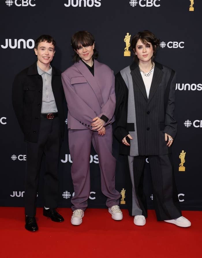 Three individuals pose together, each dressed in formal attire, with a backdrop featuring logos for JUNOs and CBC. They stand on a surface that has additional logos