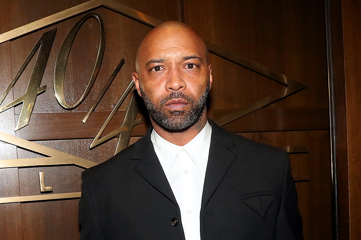 Joe Budden in a black suit with a lapel pin, standing before a wooden backdrop with '40/40 Club' etched in it