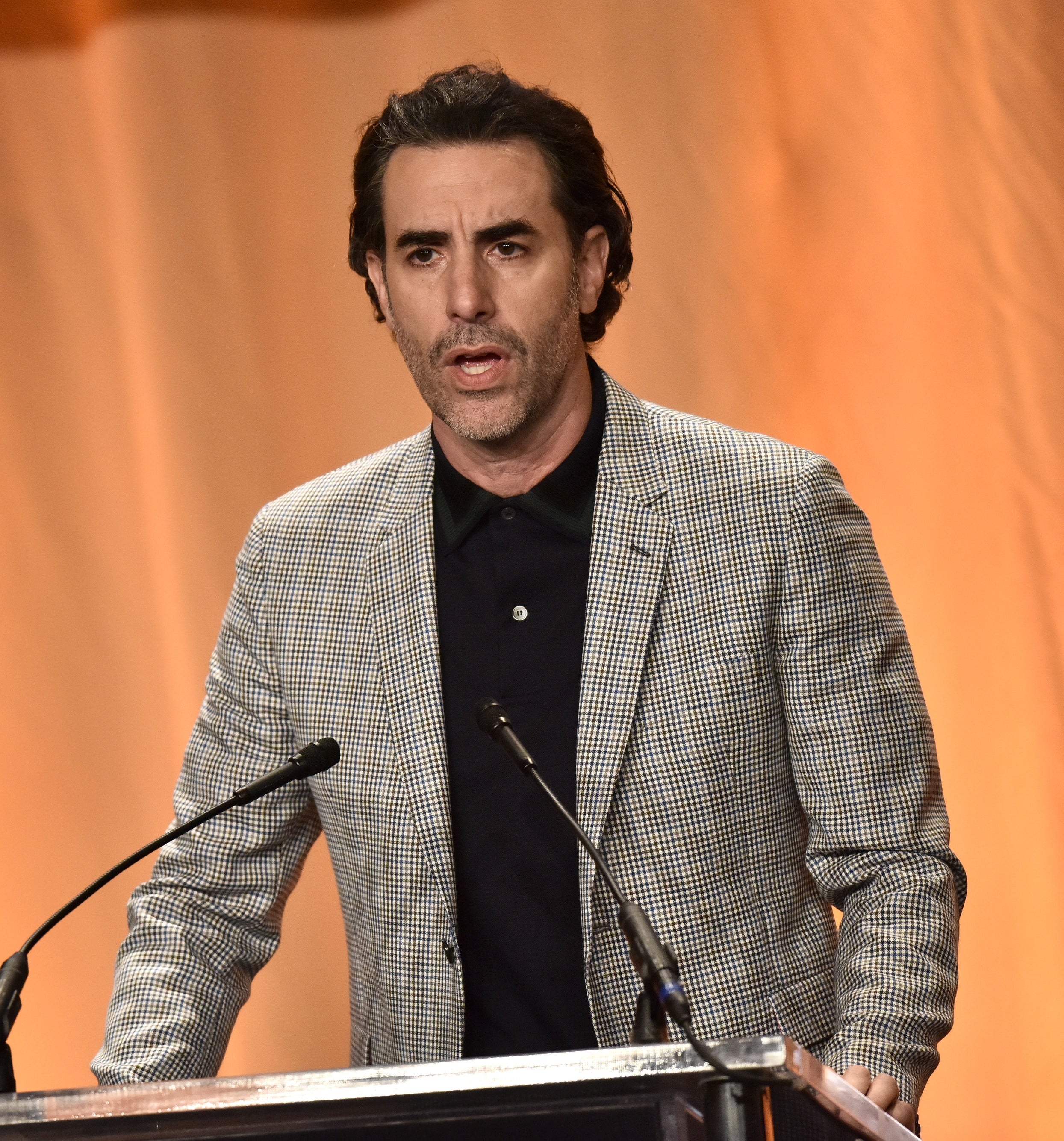 Sacha in a checkered suit and black shirt speaking at a podium