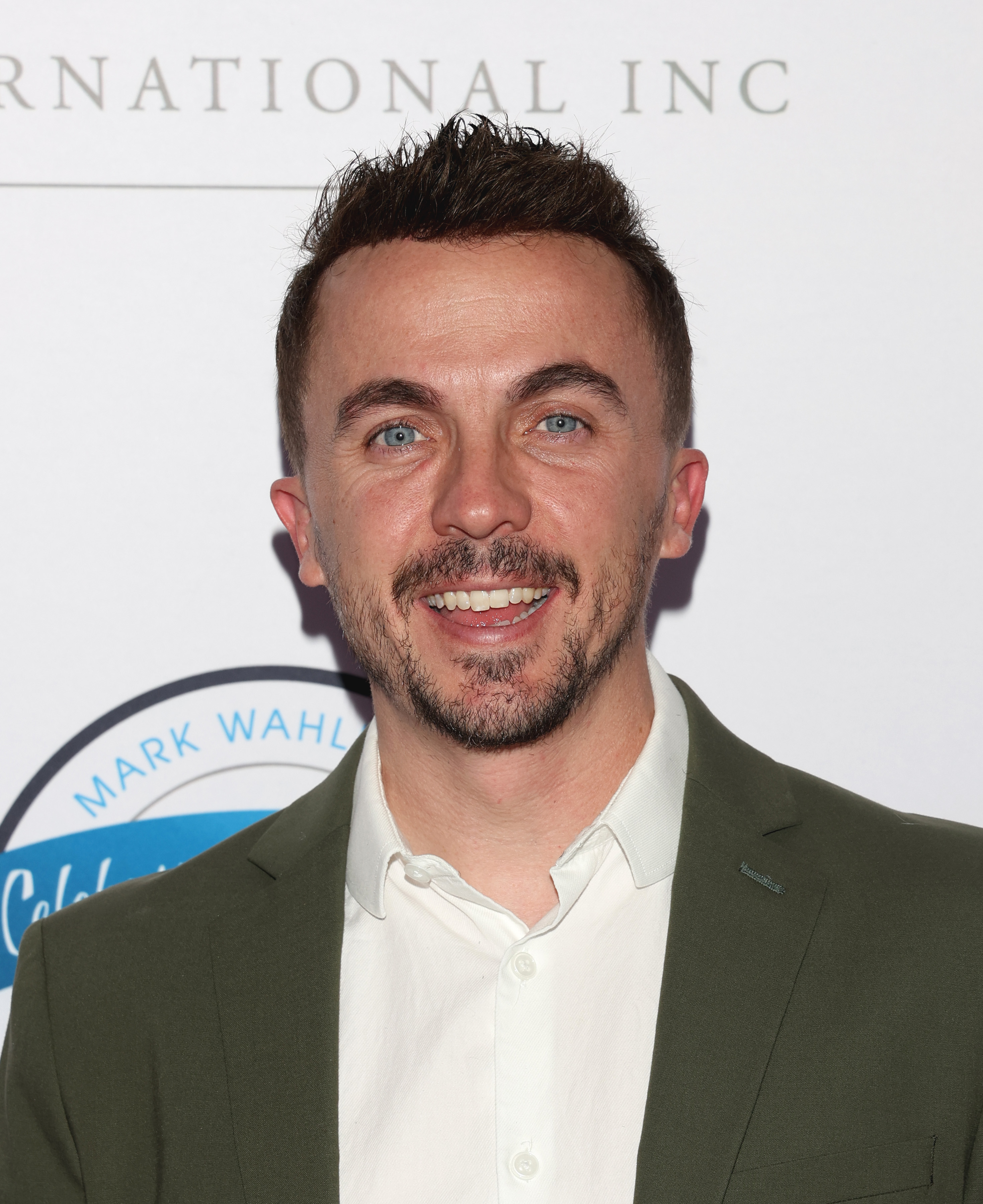 Frankie Muniz smiling in a green jacket and white shirt at a Mark Wahlberg event