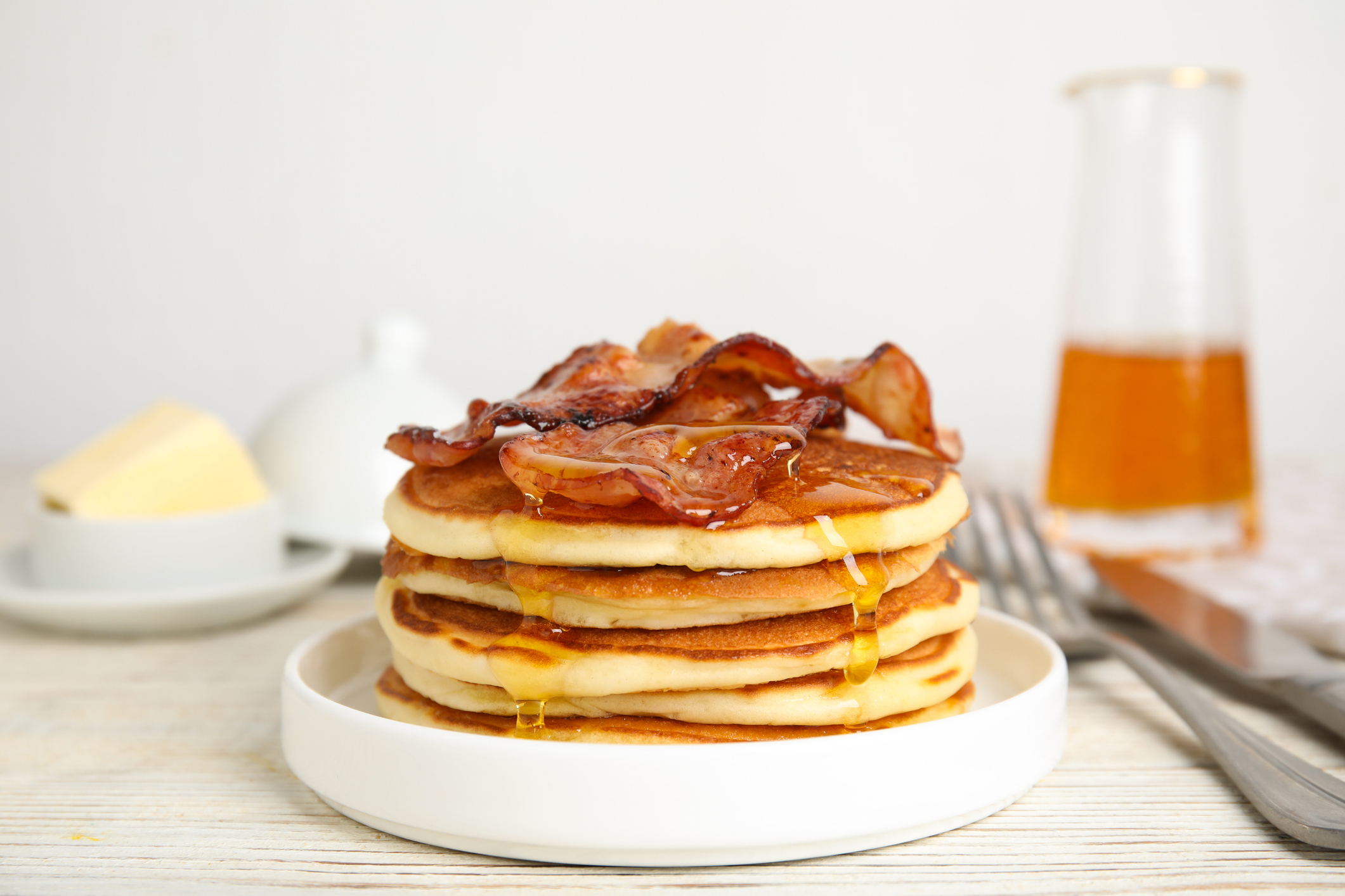 Stack of pancakes with syrup and bacon on top, beside butter and a syrup pitcher
