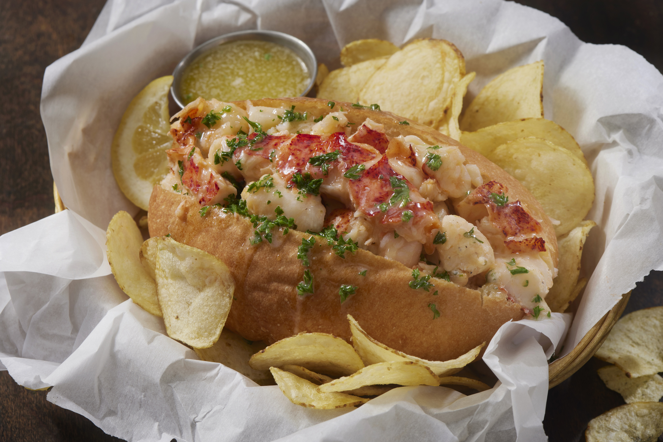 Lobster roll in bun with side of chips and lemon slices, presented in a basket