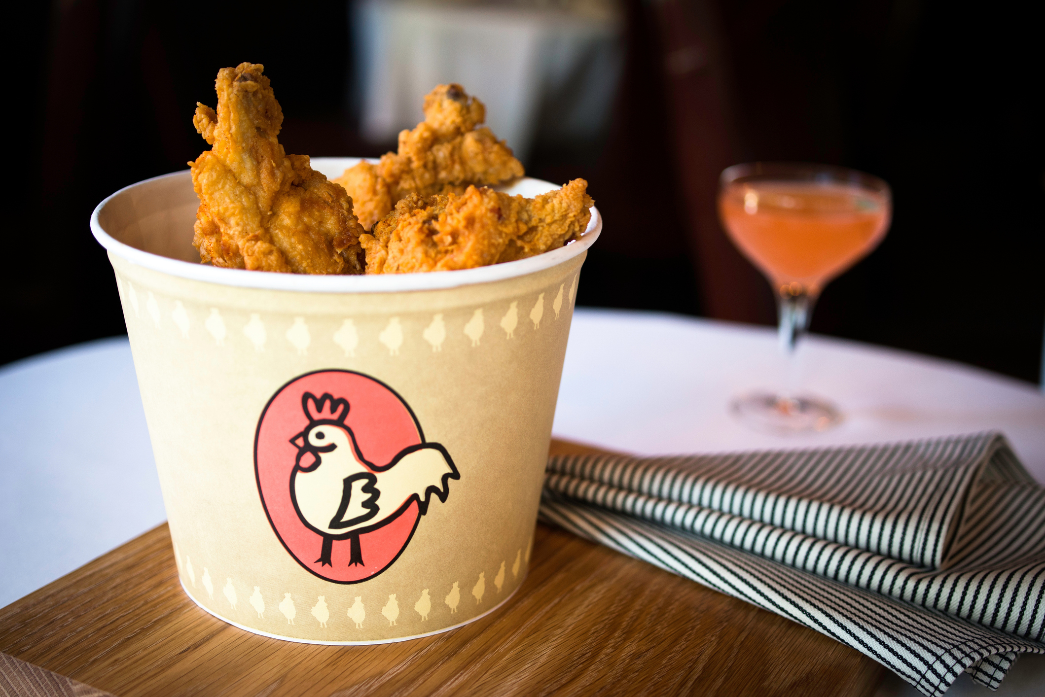 Bucket of fried chicken with a rooster logo, placed on a table with a striped napkin and a drink in the background