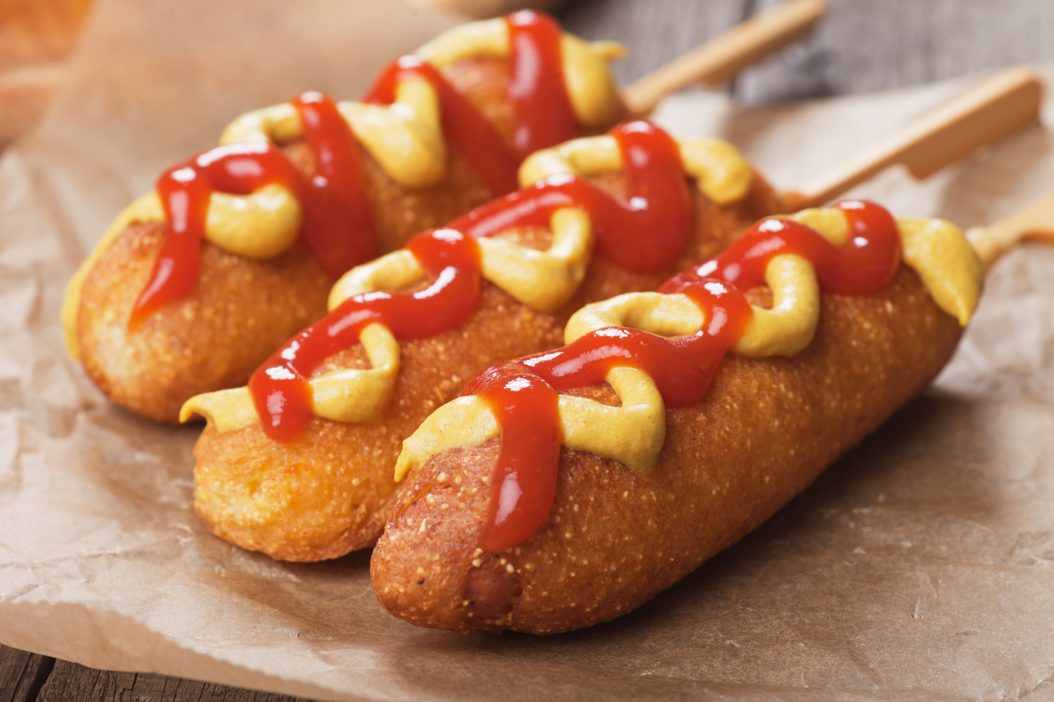 Three corn dogs with mustard and ketchup drizzled on top, displayed on parchment paper