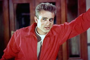 James Dean posing with one arm raised, wearing a red jacket in a still from a classic film