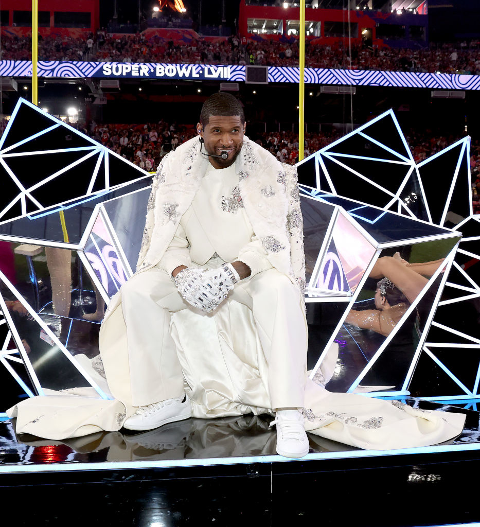 Usher onstage at the Super Bowl wearing an embellished white suit and cape, sitting with microphone in hand
