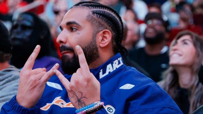 Drake sporting braids and a jacket while making a hand gesture at a basketball game