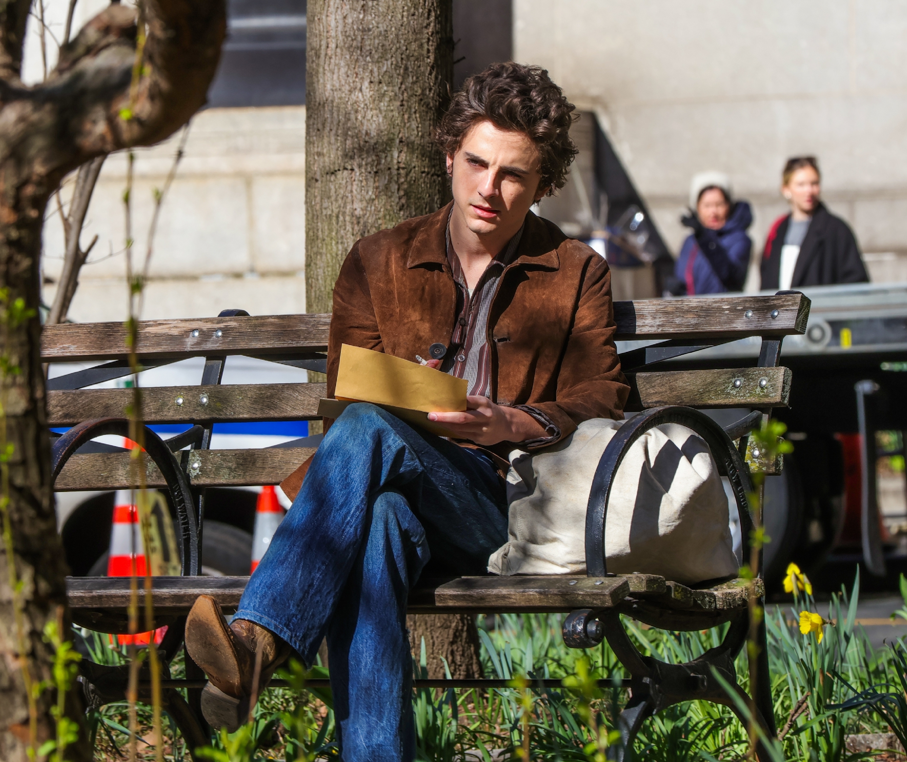 Timmy sitting on a bench with paper and pen, wearing a jacket and jeans, with trees and people in background