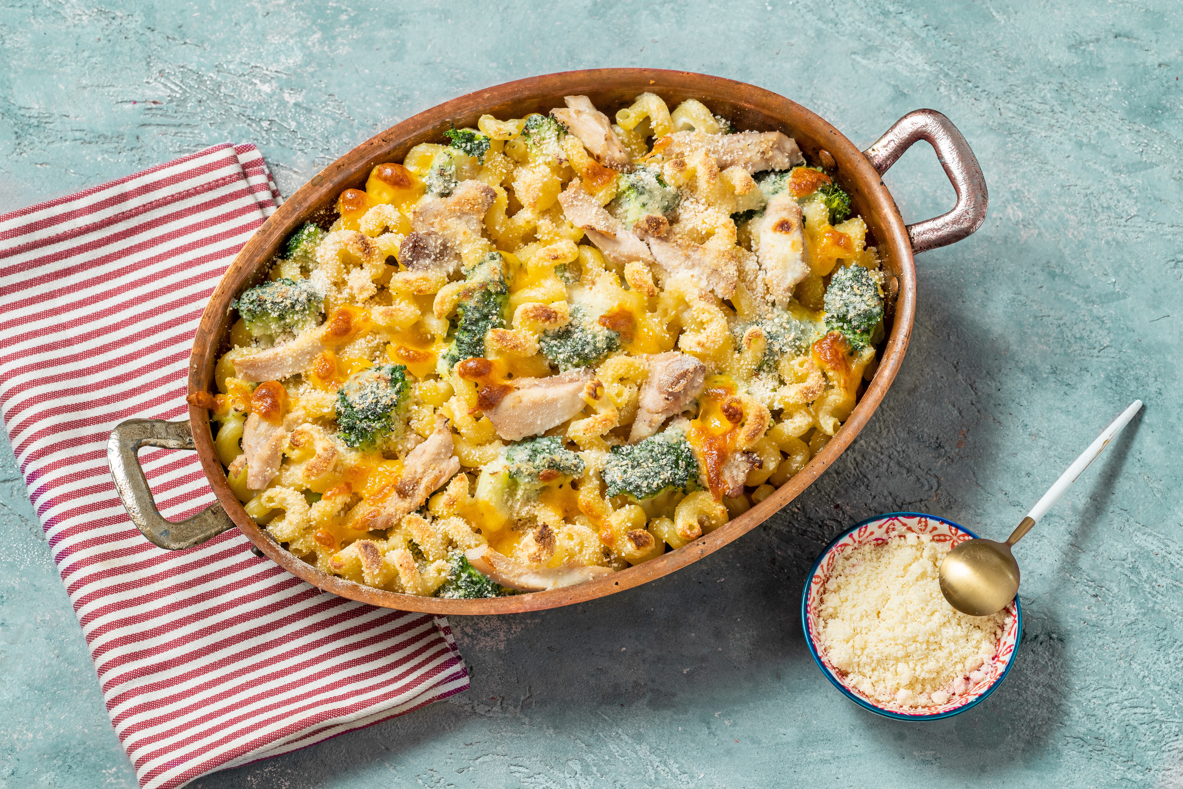 A baked chicken and broccoli pasta casserole in a dish, next to a spoonful of grated cheese