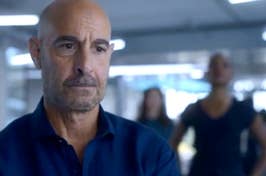 Stanley Tucci is in focus, with others in the background out of focus. He wears a blue shirt with a serious expression