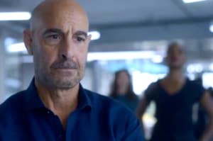 Stanley Tucci is in focus, with others in the background out of focus. He wears a blue shirt with a serious expression
