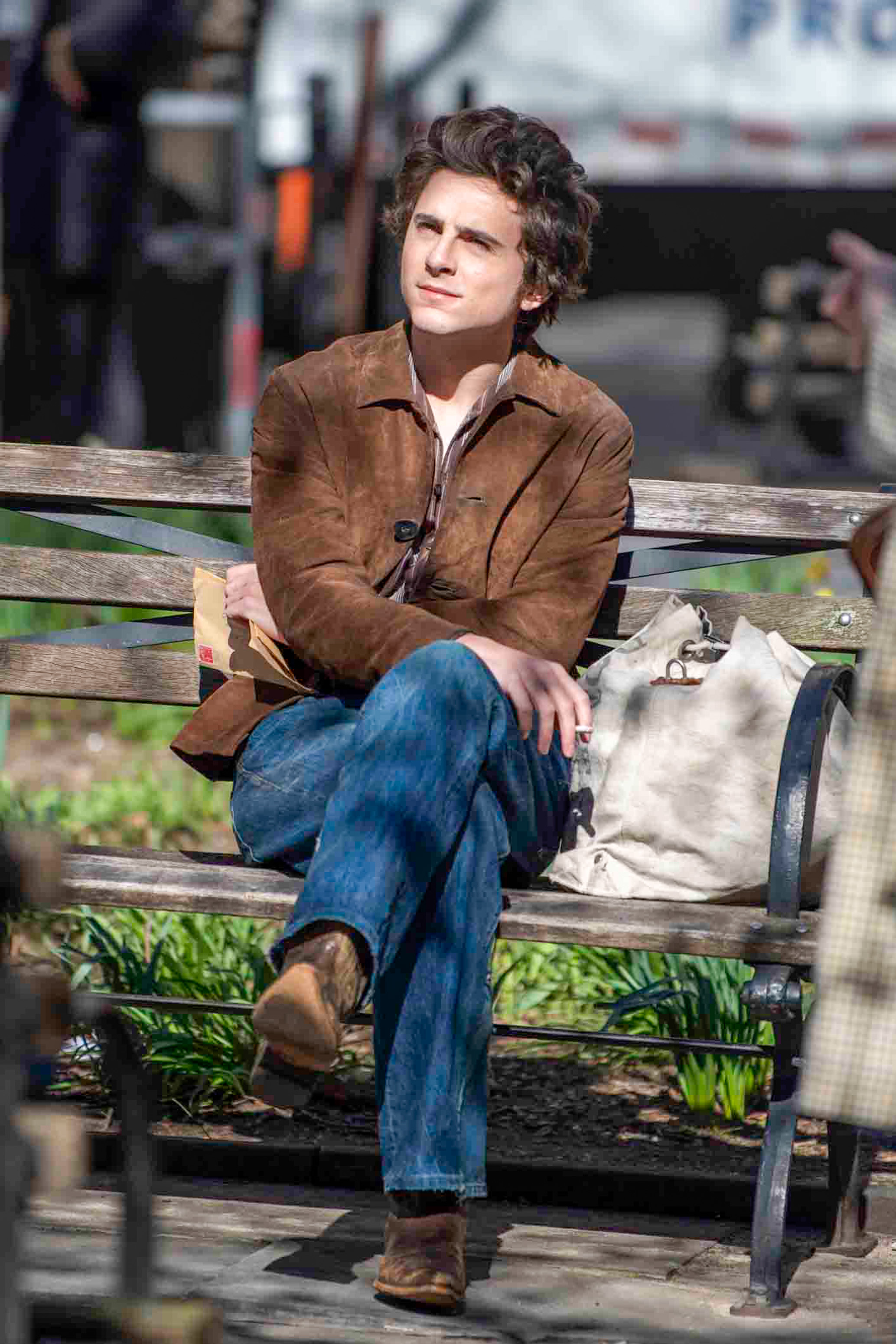 Timmy sitting on park bench with his legs crossed, wearing a brown jacket and jeans, with a tote bag beside him
