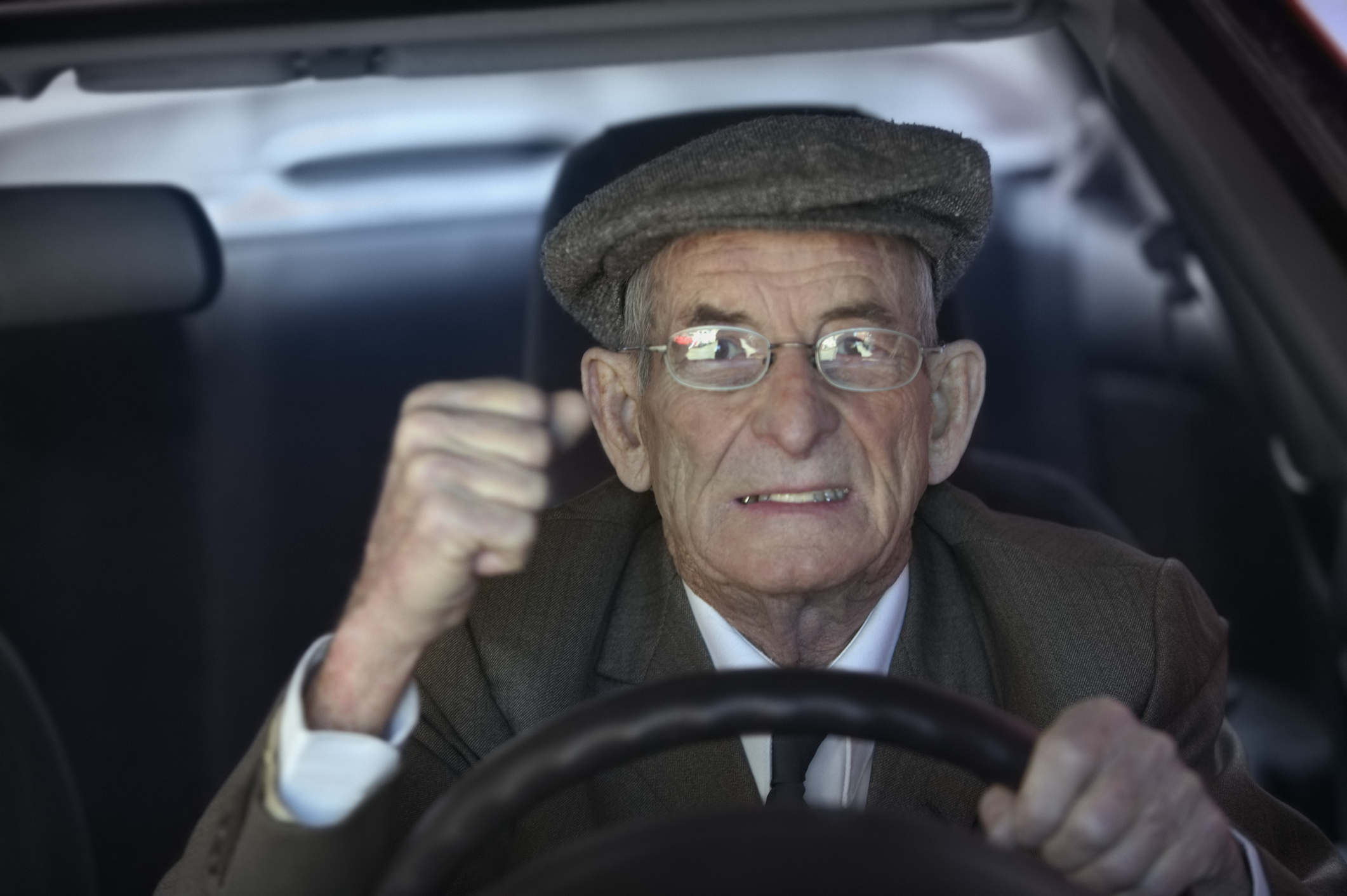 Older man in flat cap driving a car, looking forward through the windshield, his fist raised in determination
