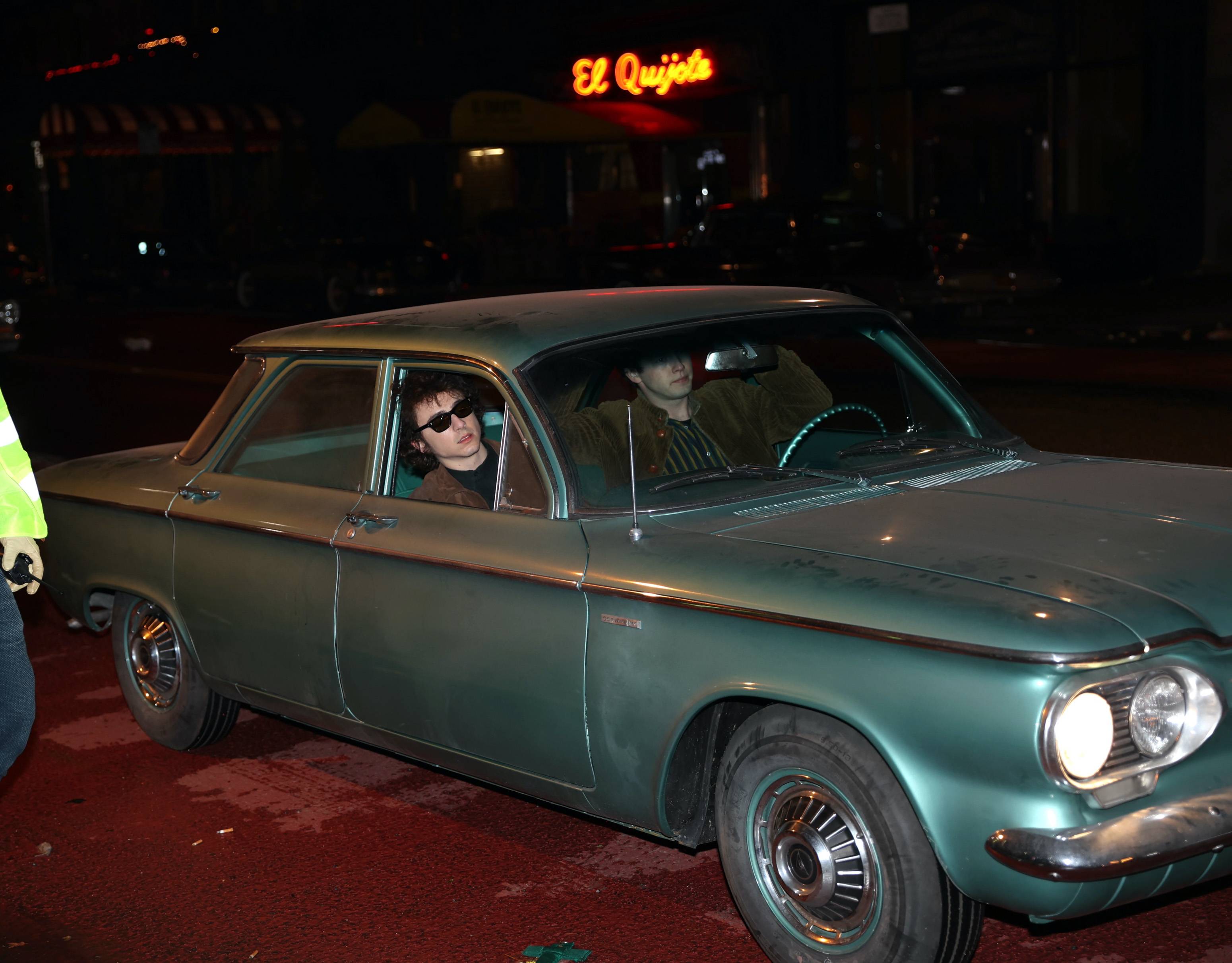Timmy wearing sunglasses with another man sitting in an old car at night with a neon sign in the background