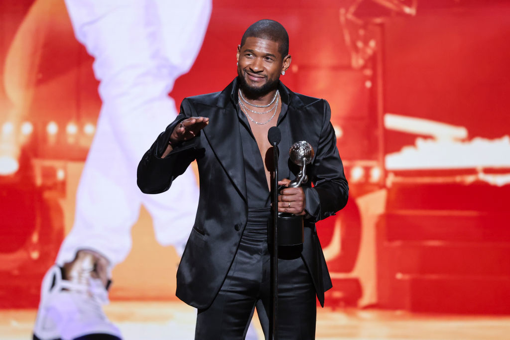 Usher onstage, wearing a black tuxedo, holding a trophy, and gesturing to the audience