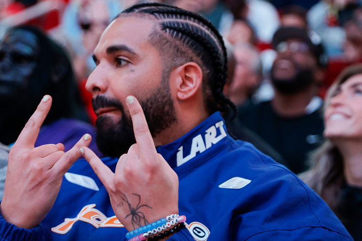 Drake sporting braids and a jacket while making a hand gesture at a basketball game