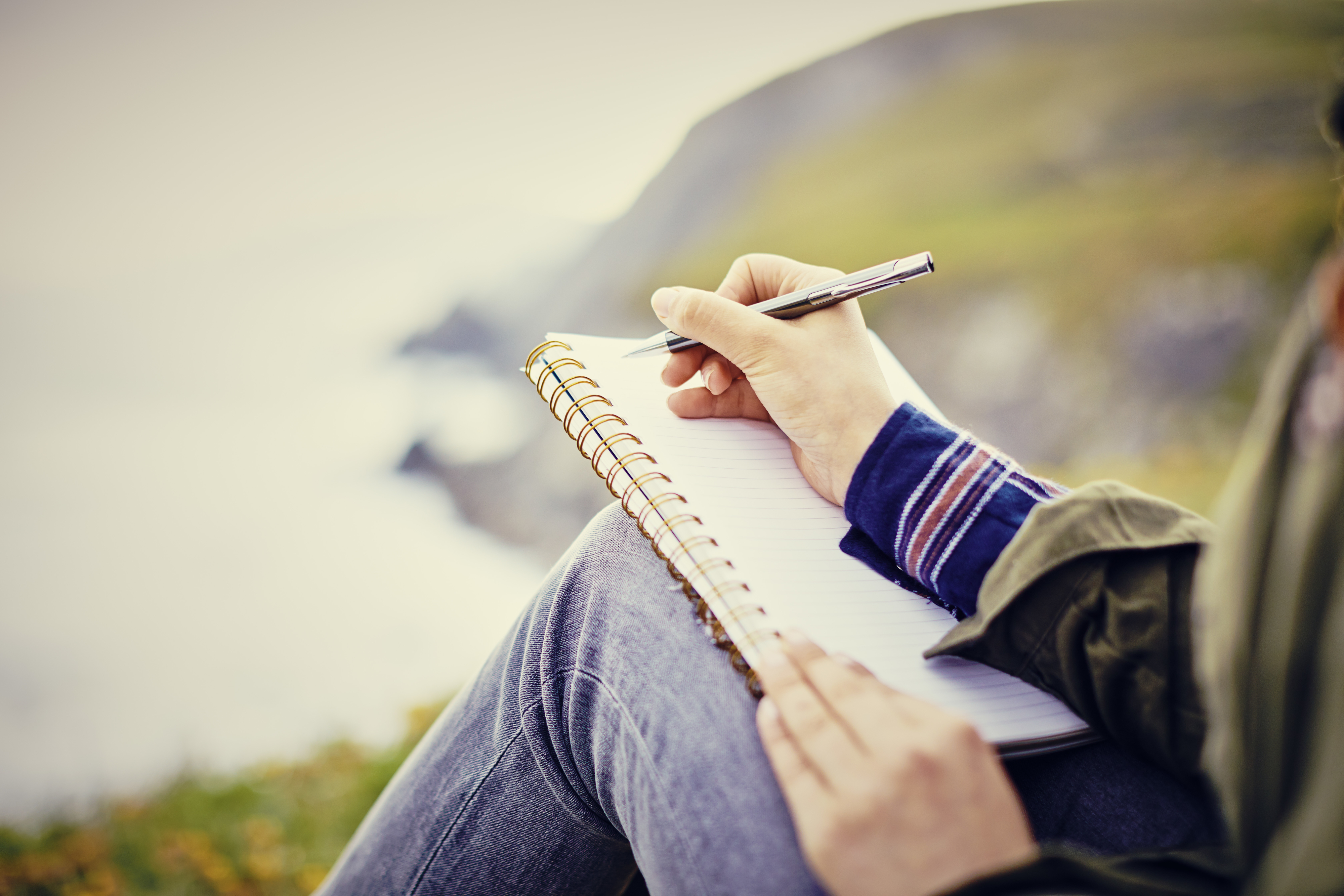 Person sitting outside writing in a spiral notebook with a pen, with nature in the background