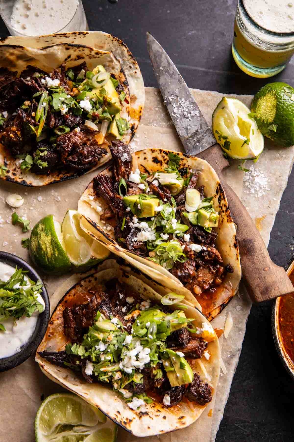 Three tacos with meat, garnished with cheese and herbs, presented on parchment paper next to limes and a knife
