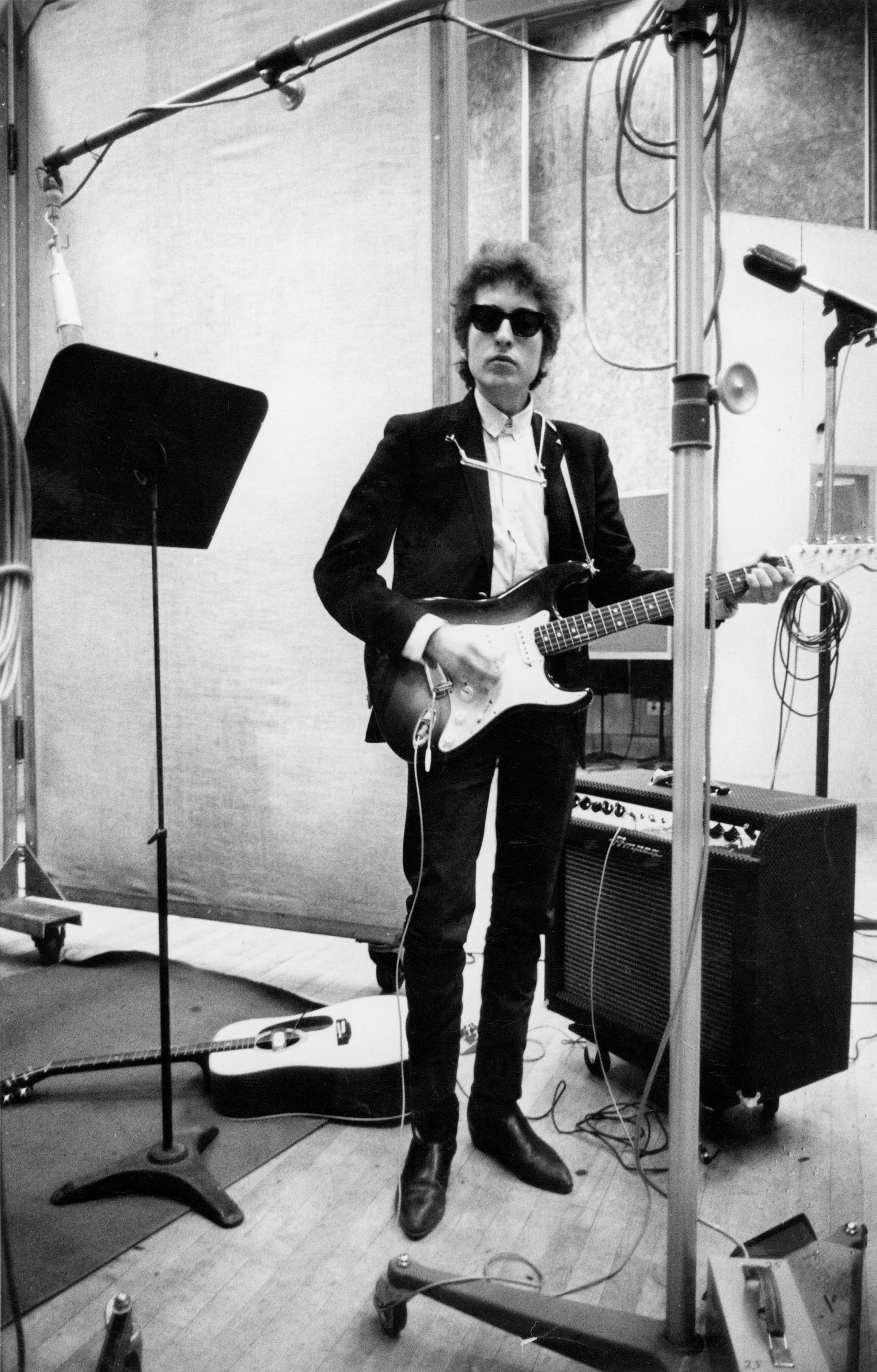 Bob in a studio with guitar, standing mic, and amp, wearing sunglasses, jacket, shirt, and straight pants