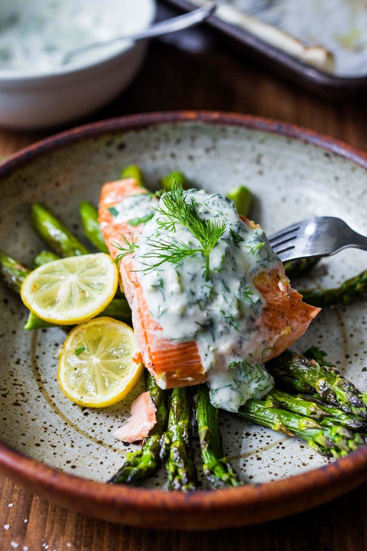 Grilled salmon with asparagus topped with dill sauce and lemon slices on a plate with a fork