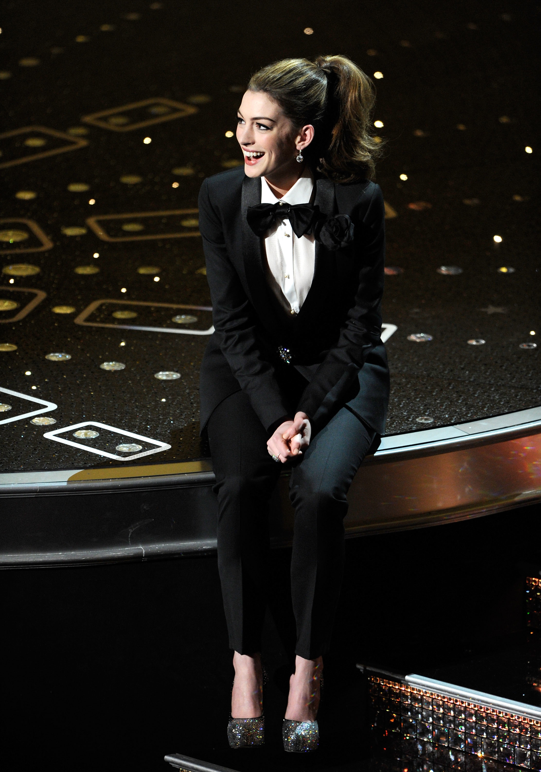 Anne Hathaway in a black tuxedo with bow tie and sparkling shoes sitting on stage while hosting the Oscars