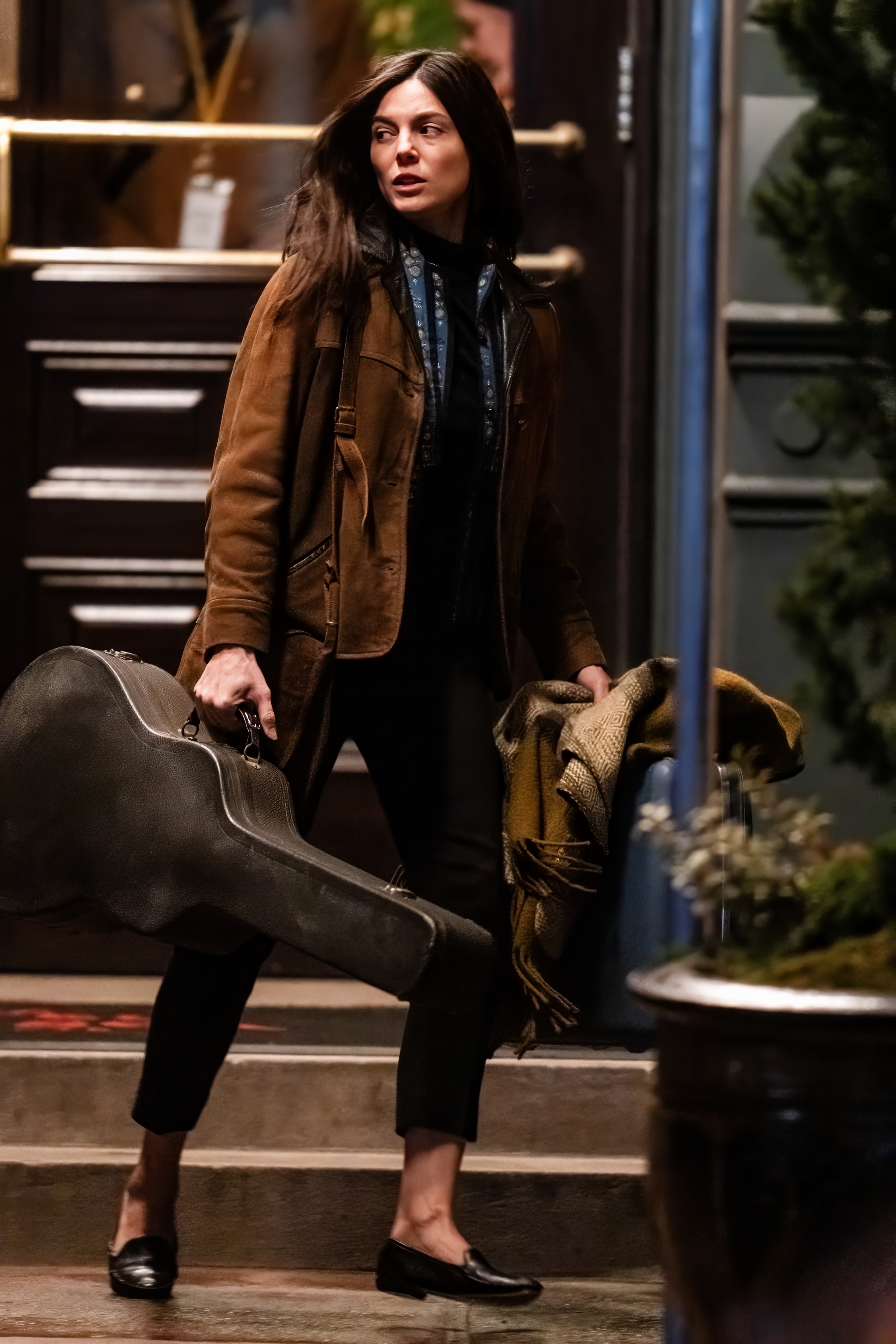 Monica, in a brown jacket, carries a guitar case on stairs
