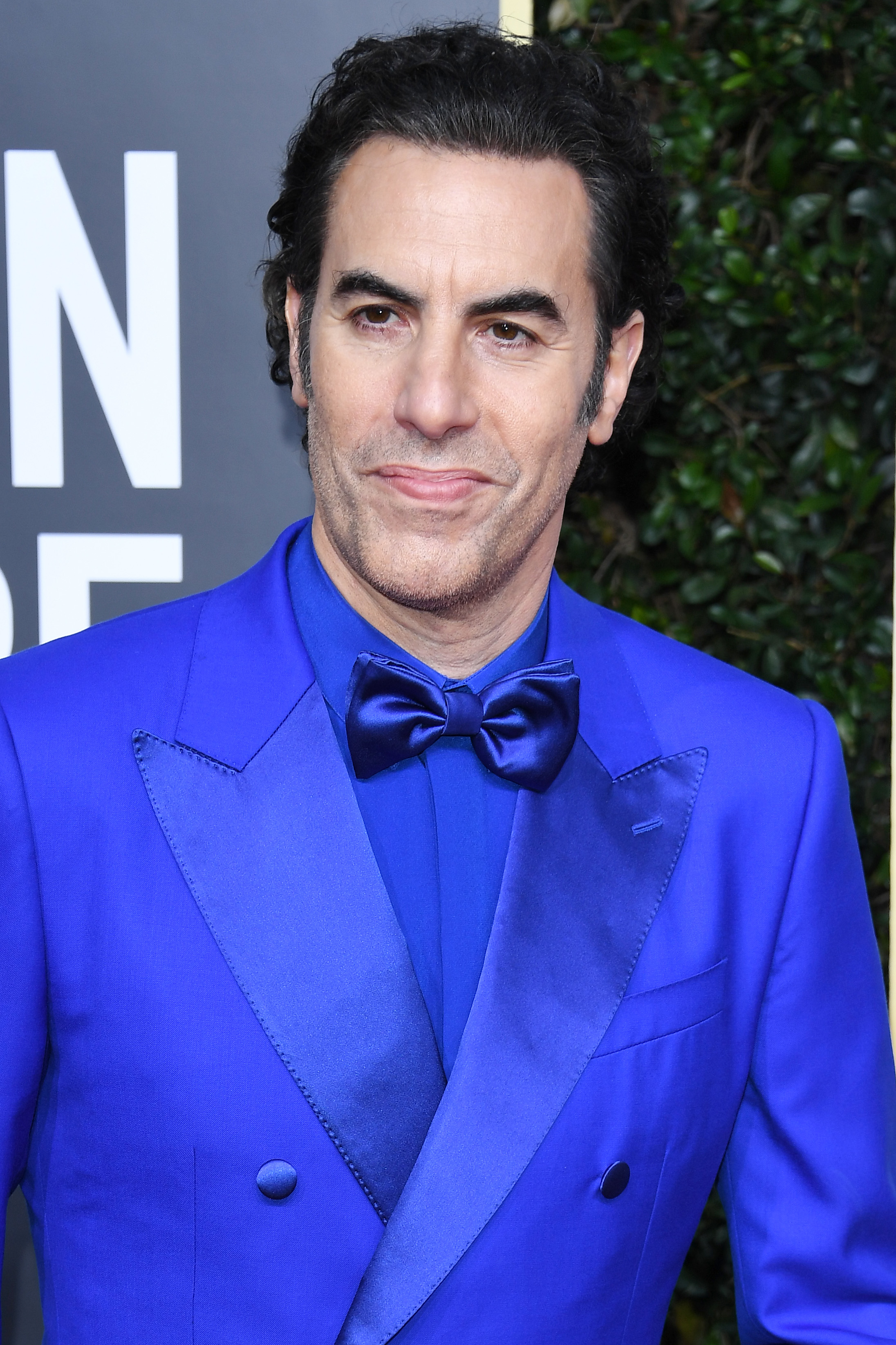 Sacha Baron Cohen in a sharp blue suit with bow tie, posing at an event