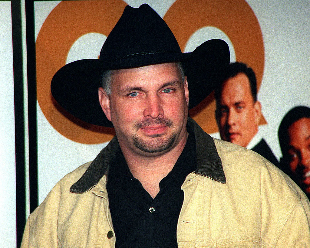 Garth Brooks wearing a cowboy hat and light jacket at an event with movie posters in the background